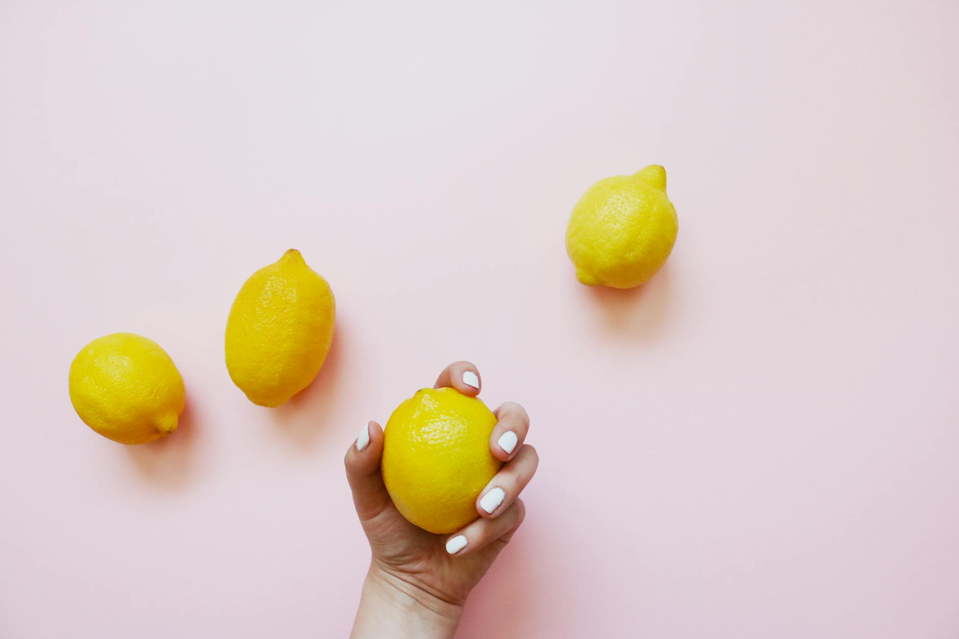 A hand holding a fresh lemon on a soft pink background with three additional lemons nearby