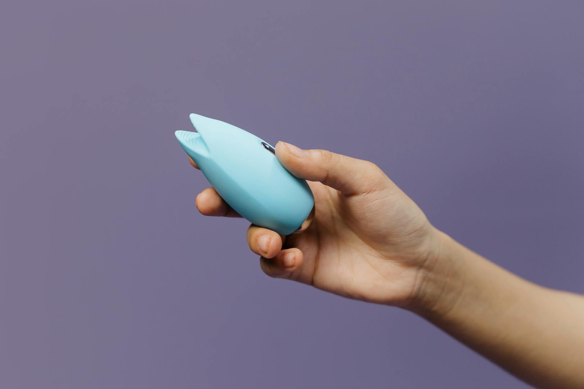 A blue silicone clitoral vibrator in hand against a solid purple background, symbolizing self-care and couples wellness